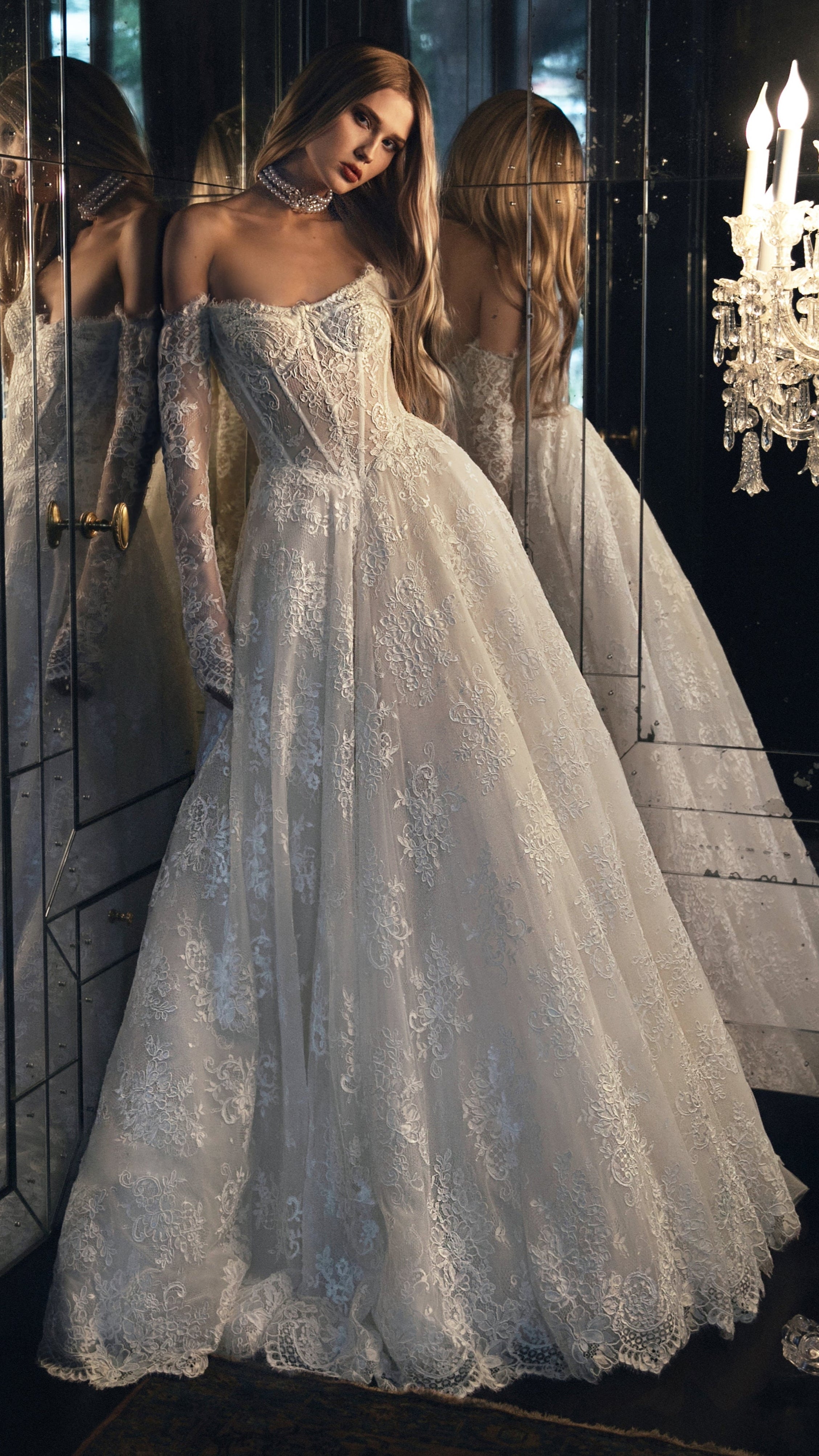 Woman in a white lace wedding dress standing in front of a mirror with chandeliers.