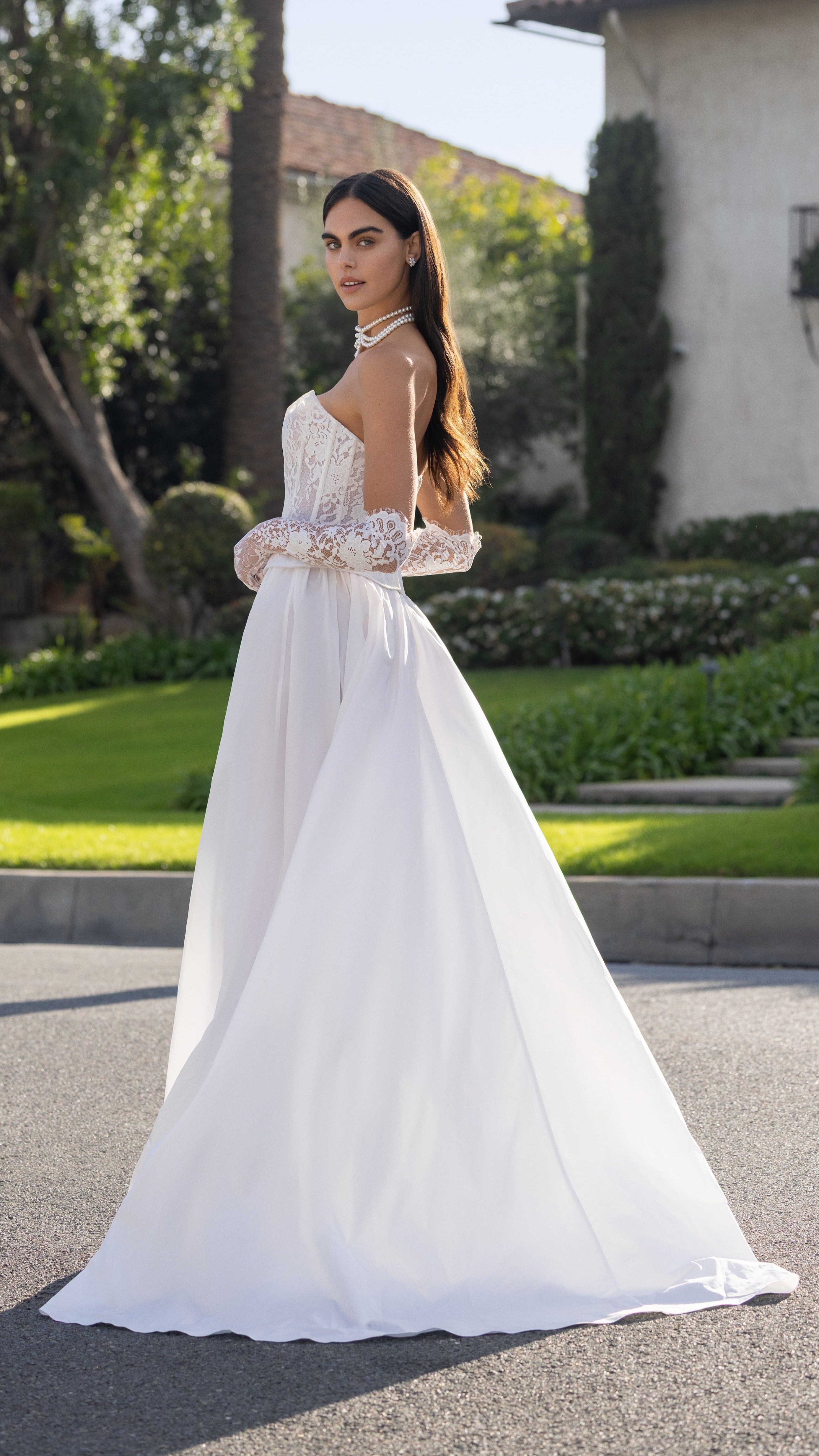 Woman in a white wedding dress standing on a street with greenery and a building in the background