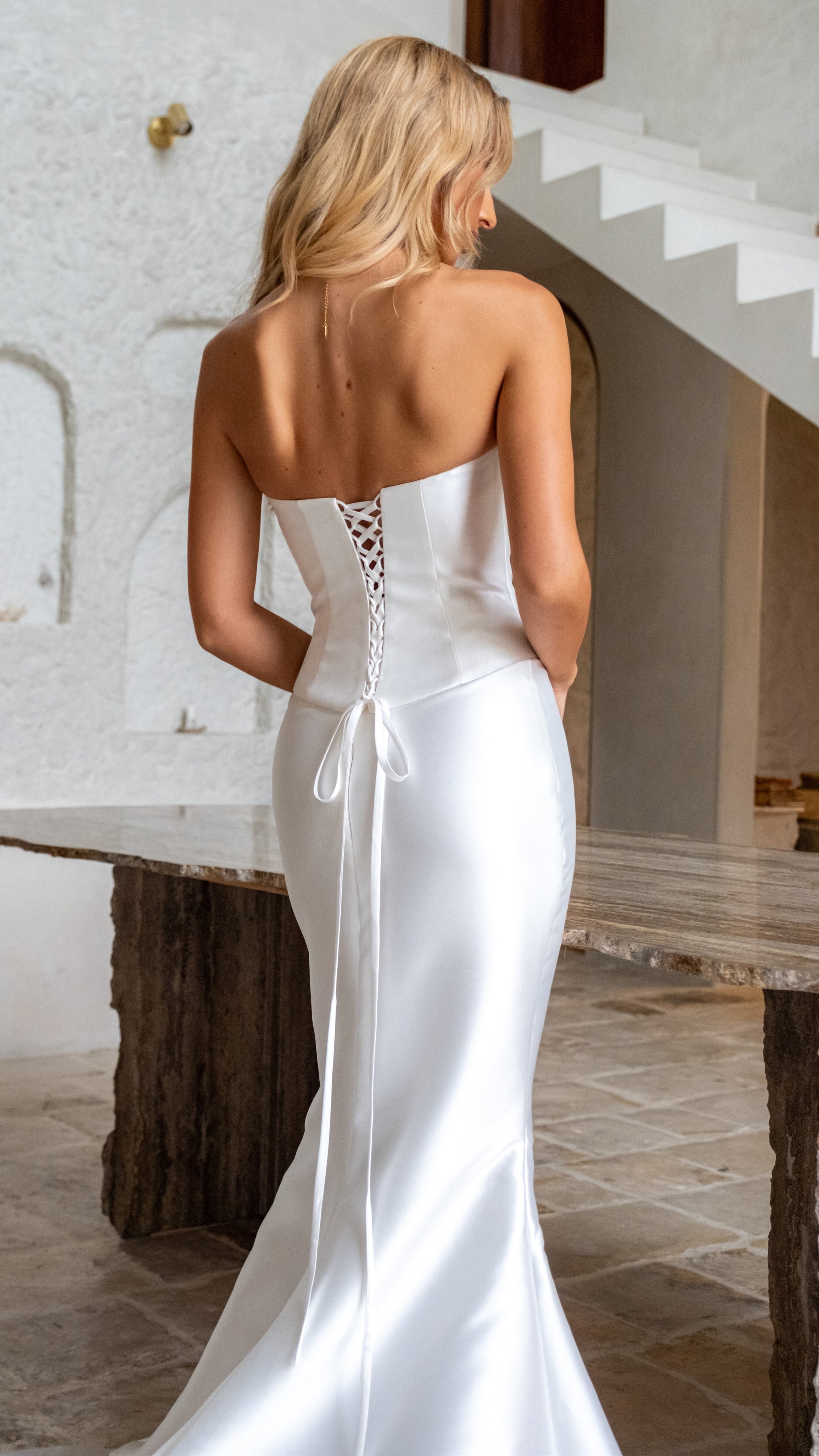 Woman in a white strapless wedding dress standing in a room with a textured wall and wooden table.