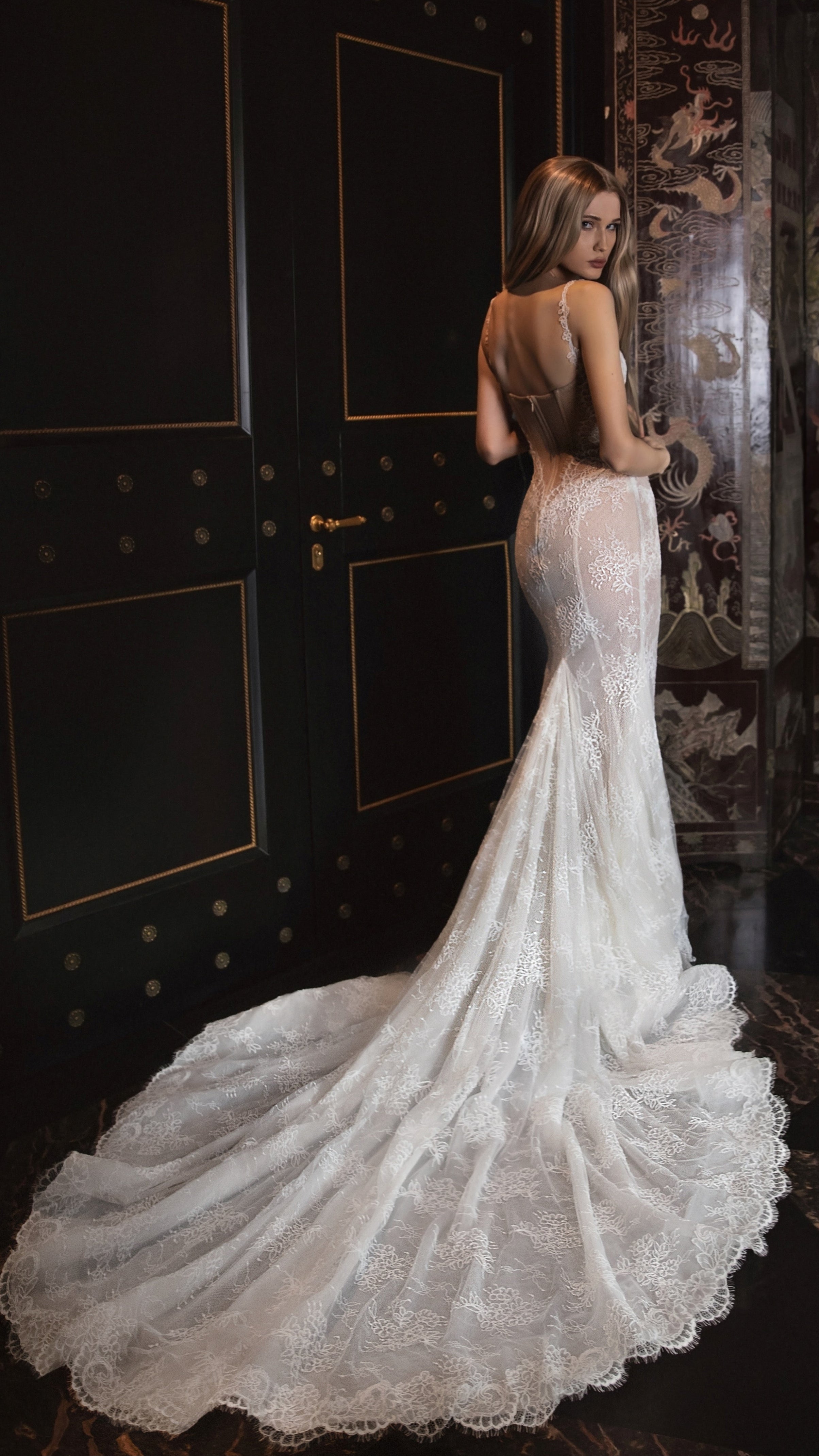 Woman in a white lace wedding dress standing in a dark room with decorative walls.