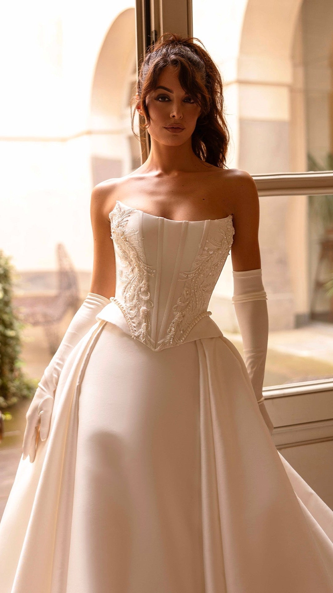 Woman in a white wedding dress standing in a sunlit room with large windows.