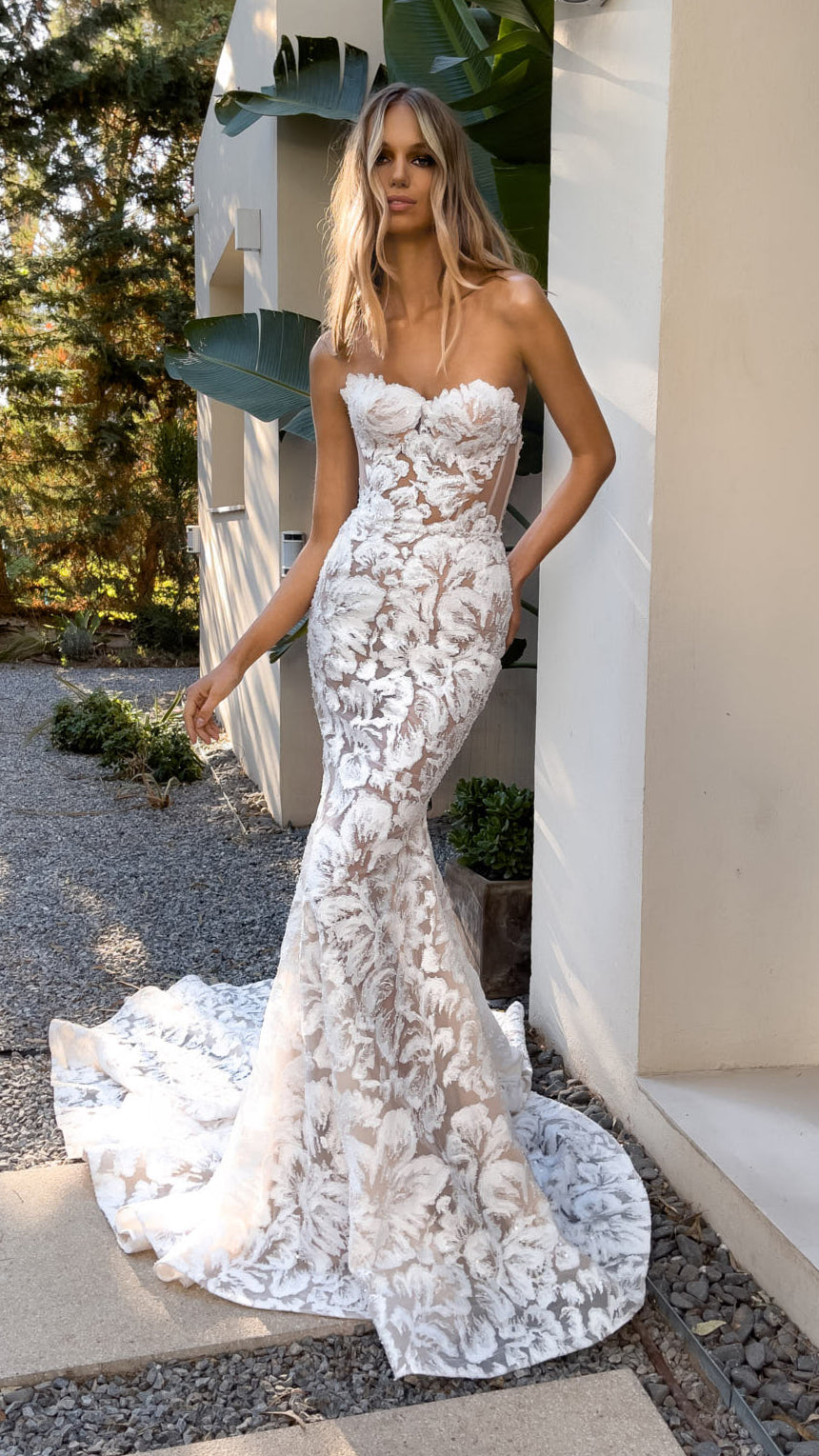 Woman in a white lace wedding dress standing outdoors near a modern building.
