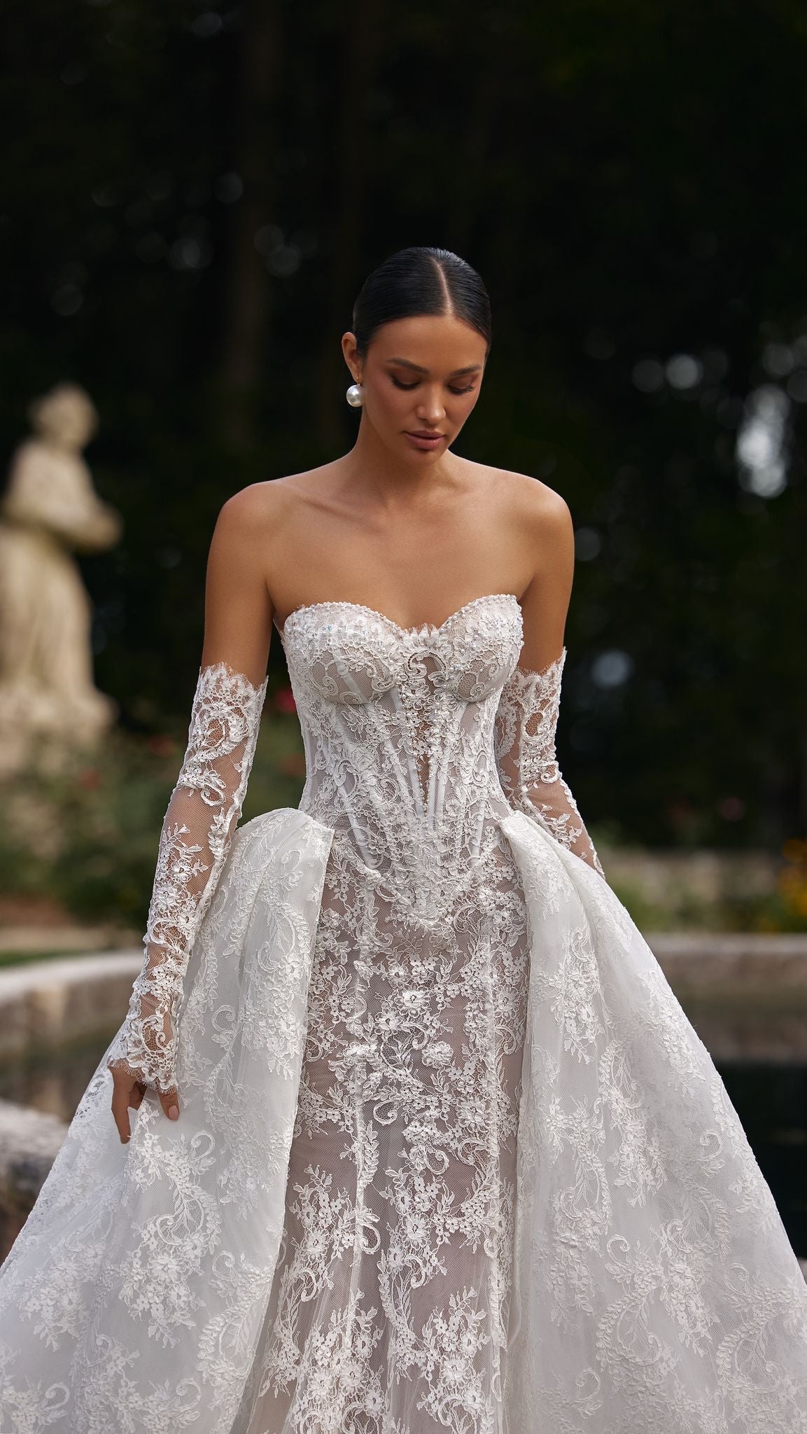 Woman in a white lace wedding dress standing in a garden setting.