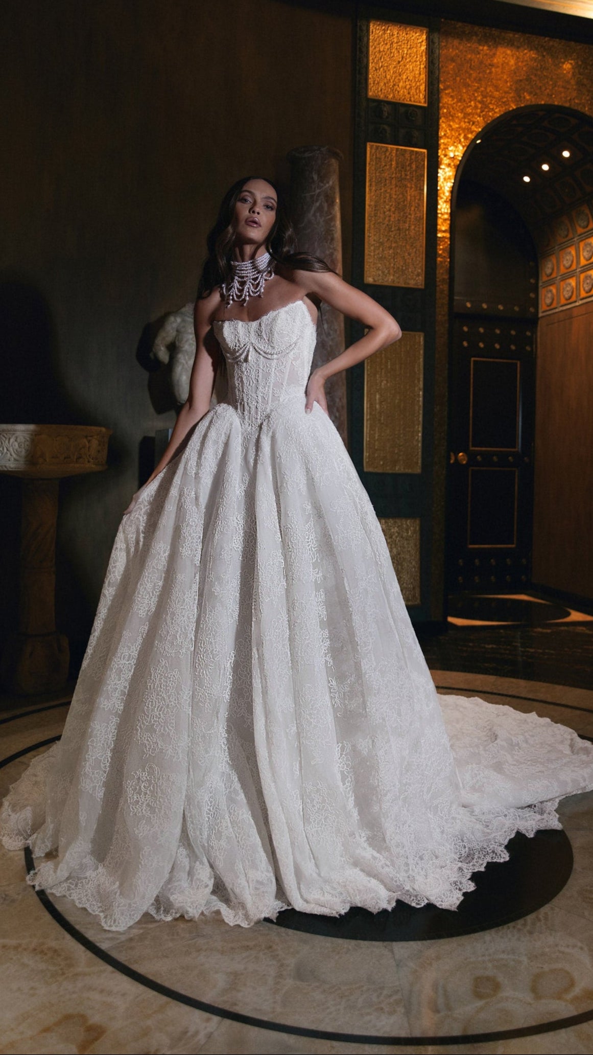 Woman in a white lace wedding dress standing in an elegant interior setting.