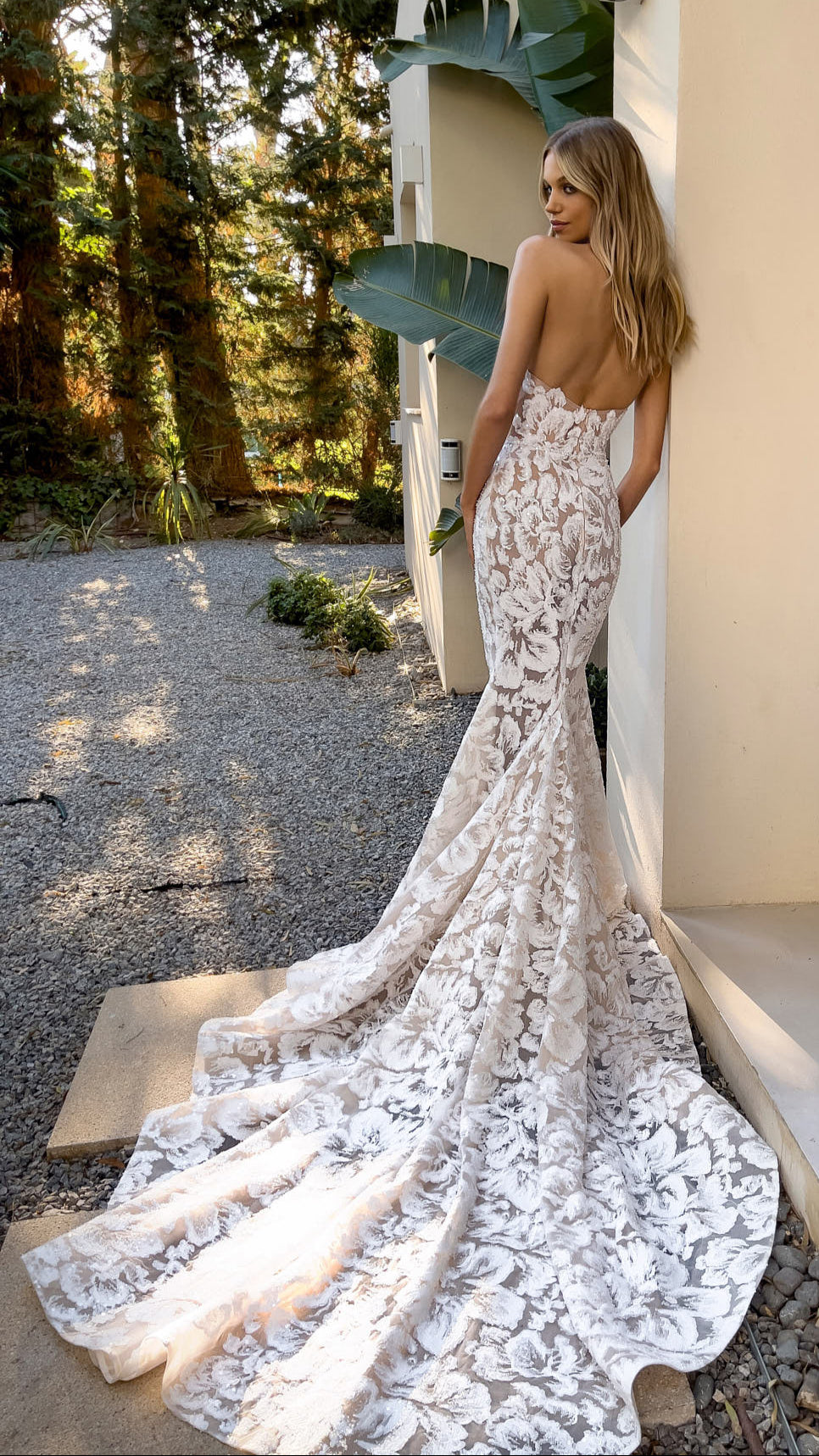 Woman in a white lace wedding dress standing outdoors near a building with plants.