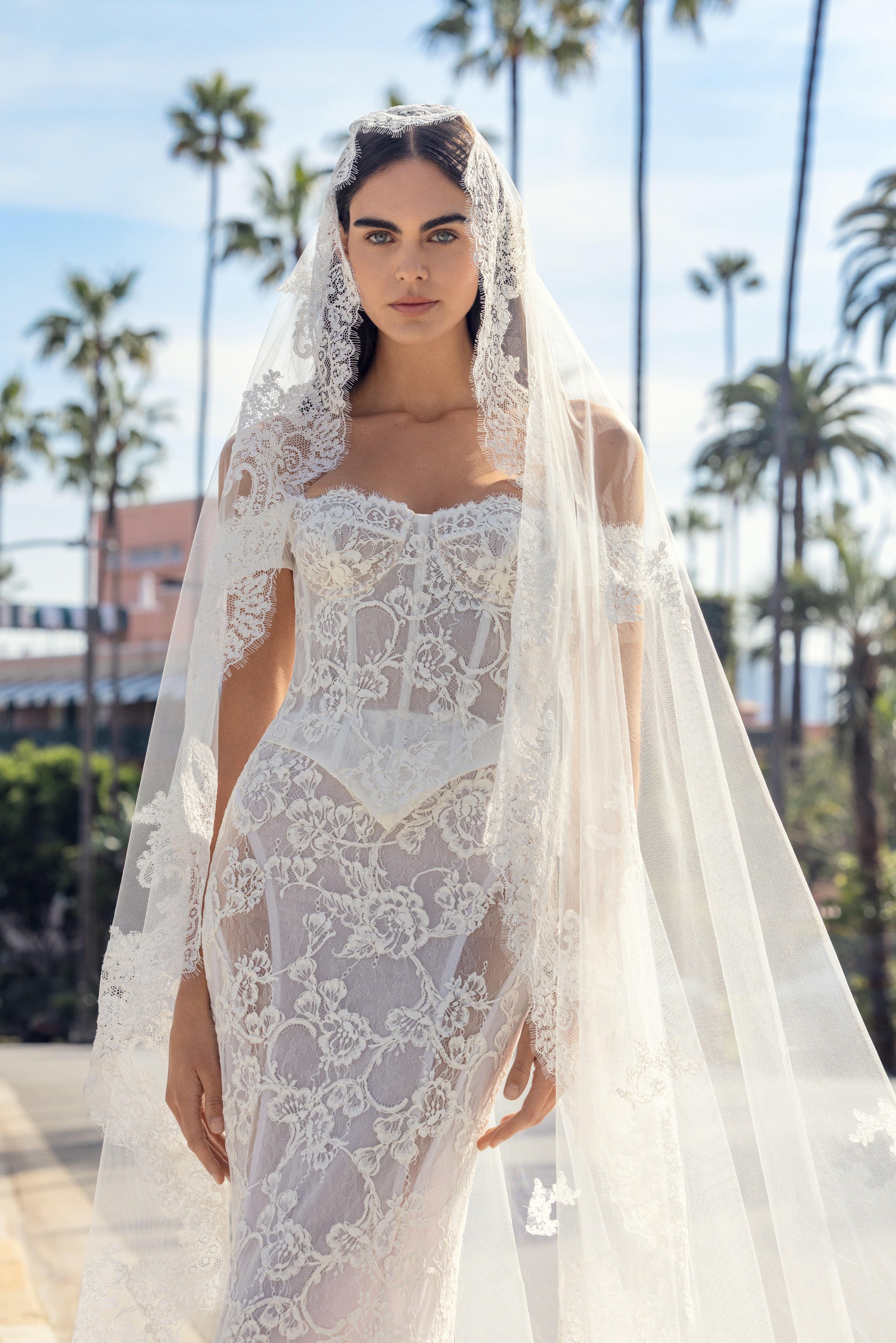 Woman wearing a white lace wedding dress with a matching veil in an outdoor setting with palm trees.