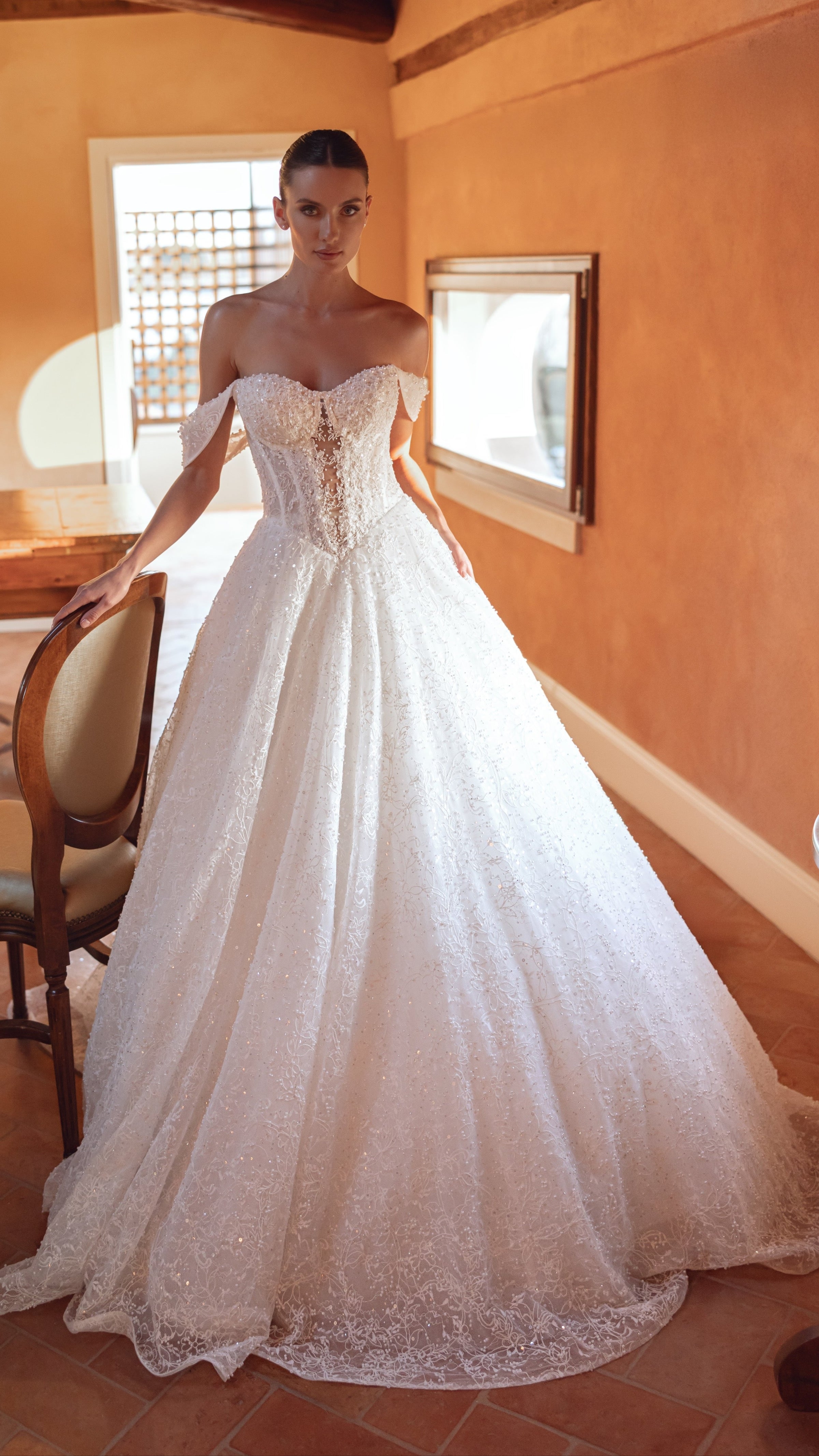 Woman in a white wedding dress standing in a rustic room with wooden beams and a chair.