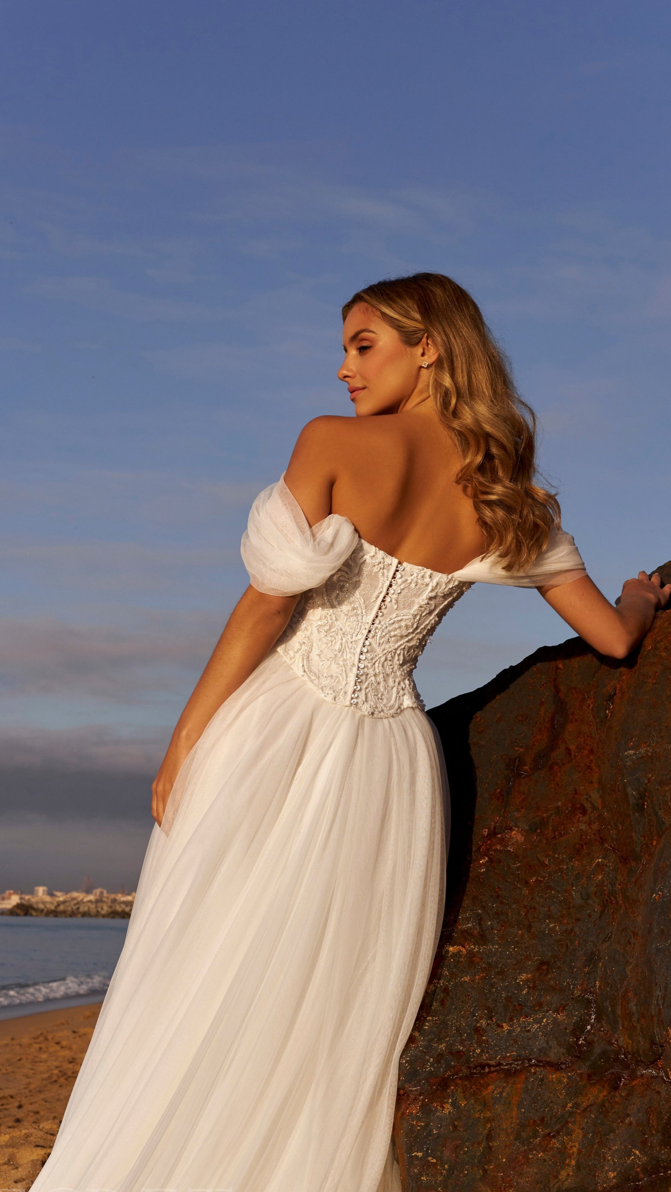 Woman in a white wedding dress standing on a rocky outcrop with a city skyline in the distance.