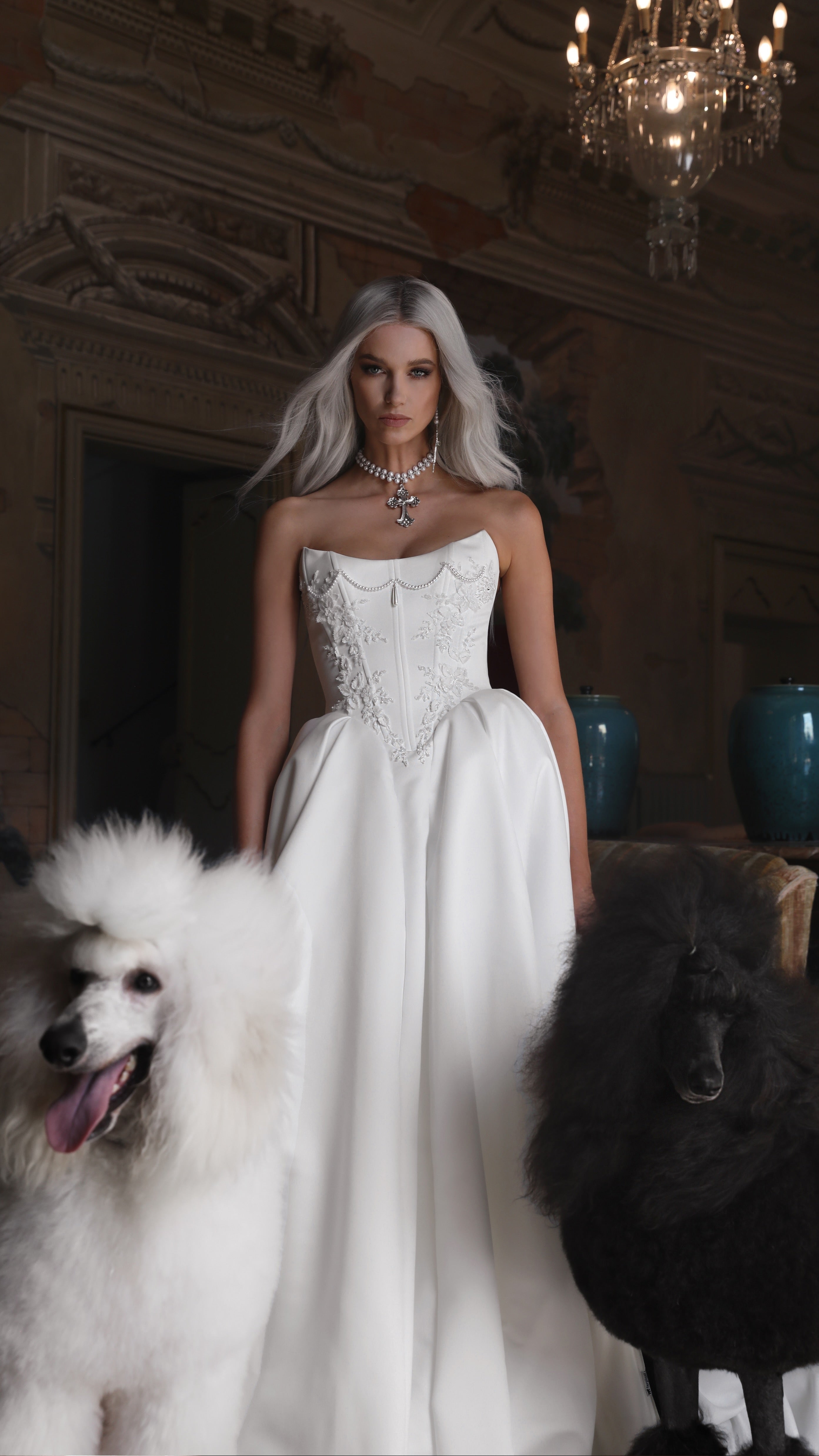 Woman in a white wedding dress standing with two dogs in an elegant room.