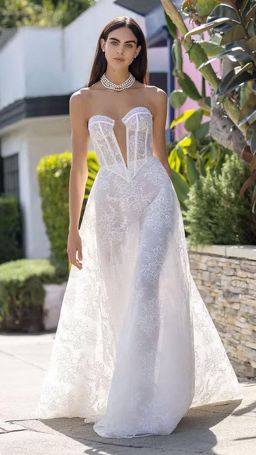Woman in a white wedding dress standing outdoors with greenery and a building in the background