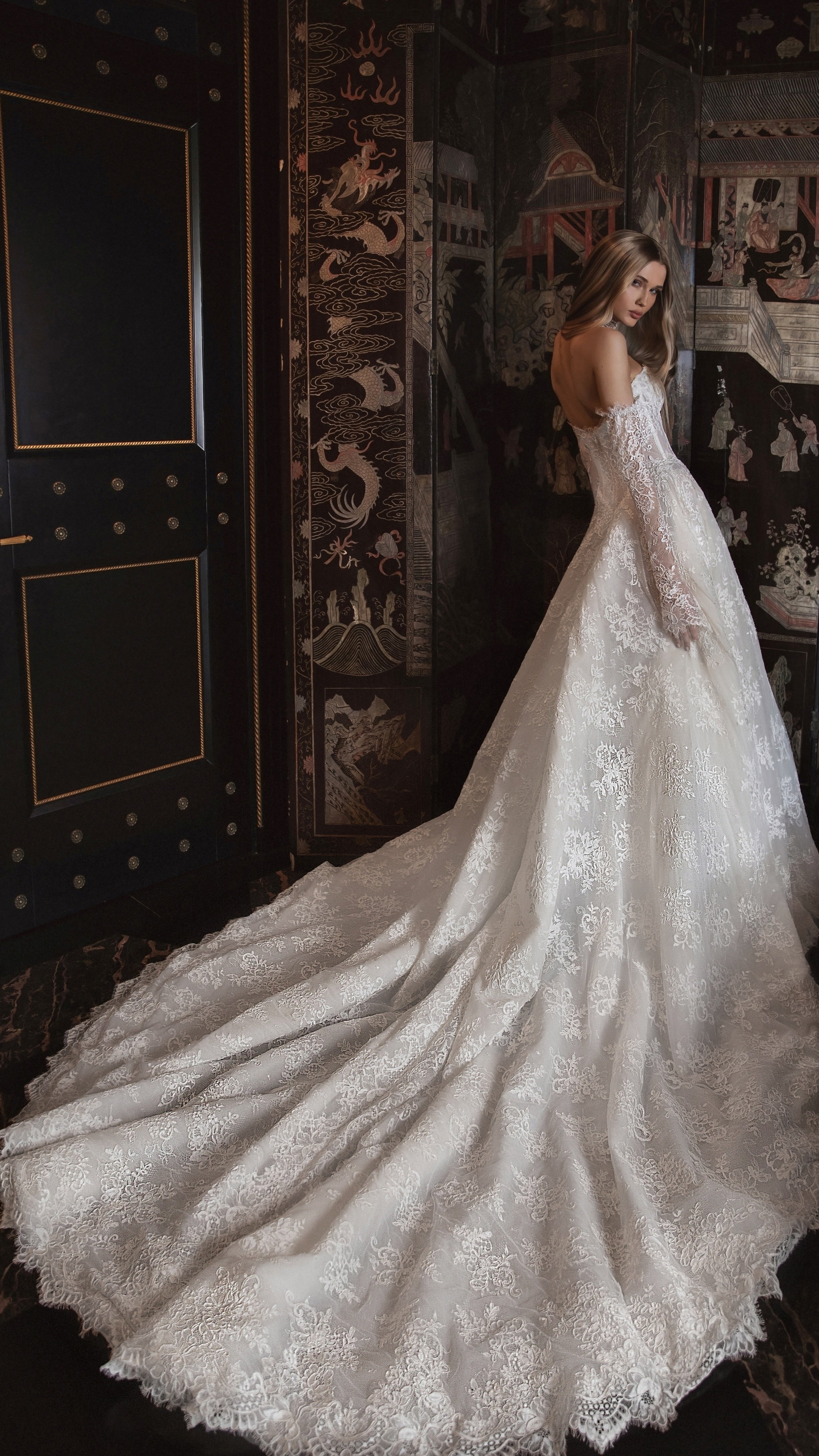 Woman in a white lace wedding dress standing in a room with decorative walls.