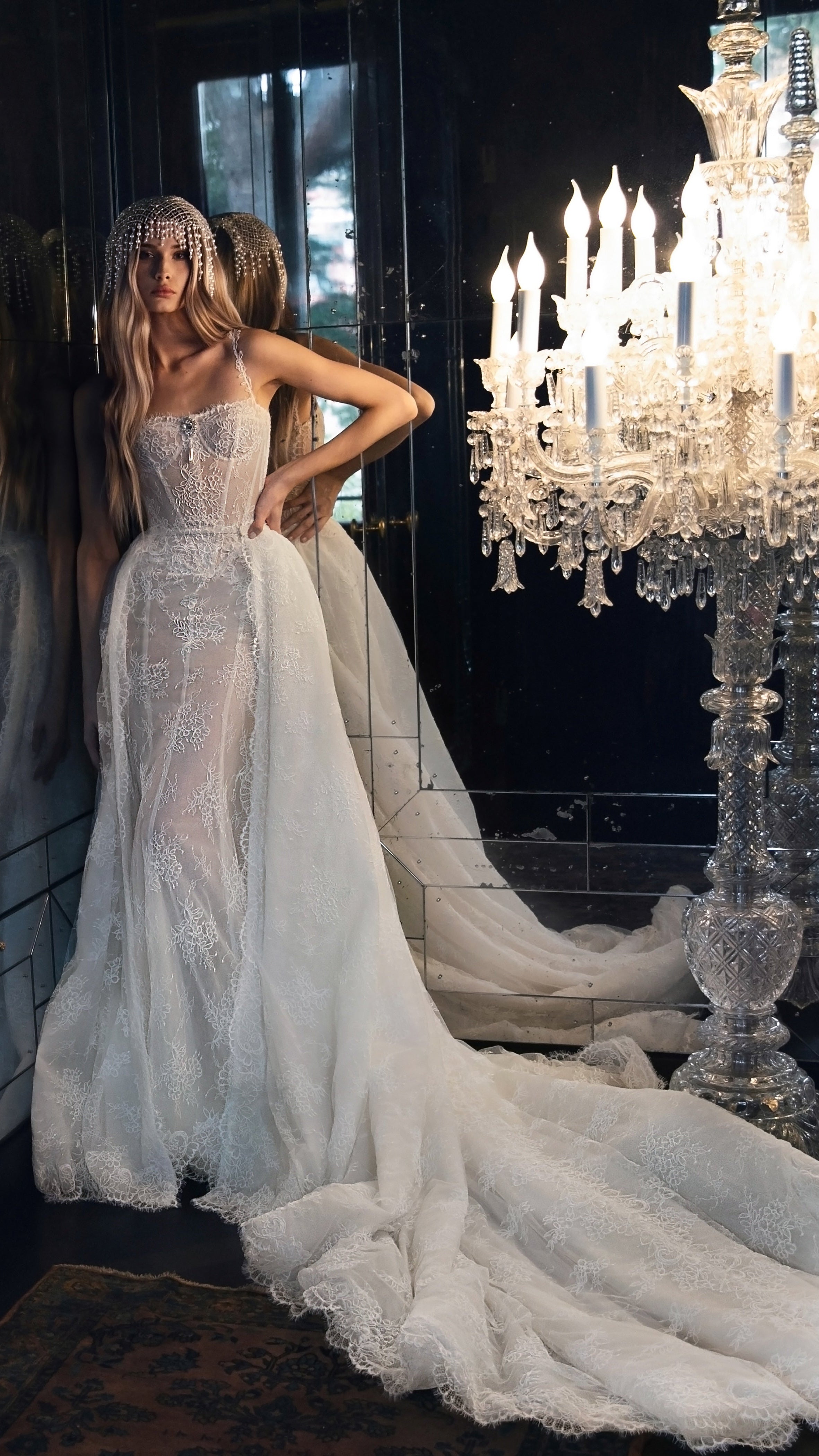 Woman in a white wedding dress standing next to a grand chandelier in an elegant room.