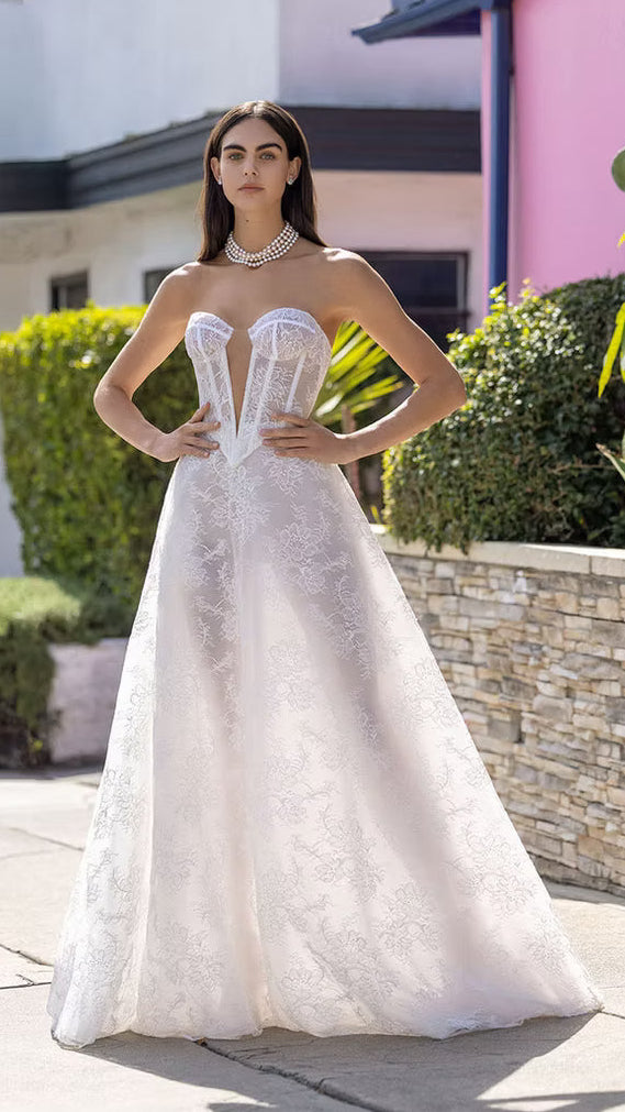 Woman in a white strapless wedding dress standing outdoors with a building and greenery in the background
