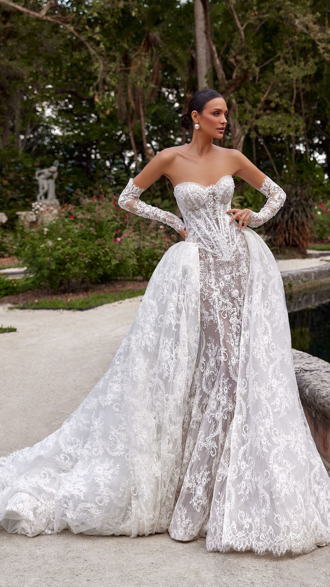 Woman in a white lace wedding dress standing outdoors with greenery in the background