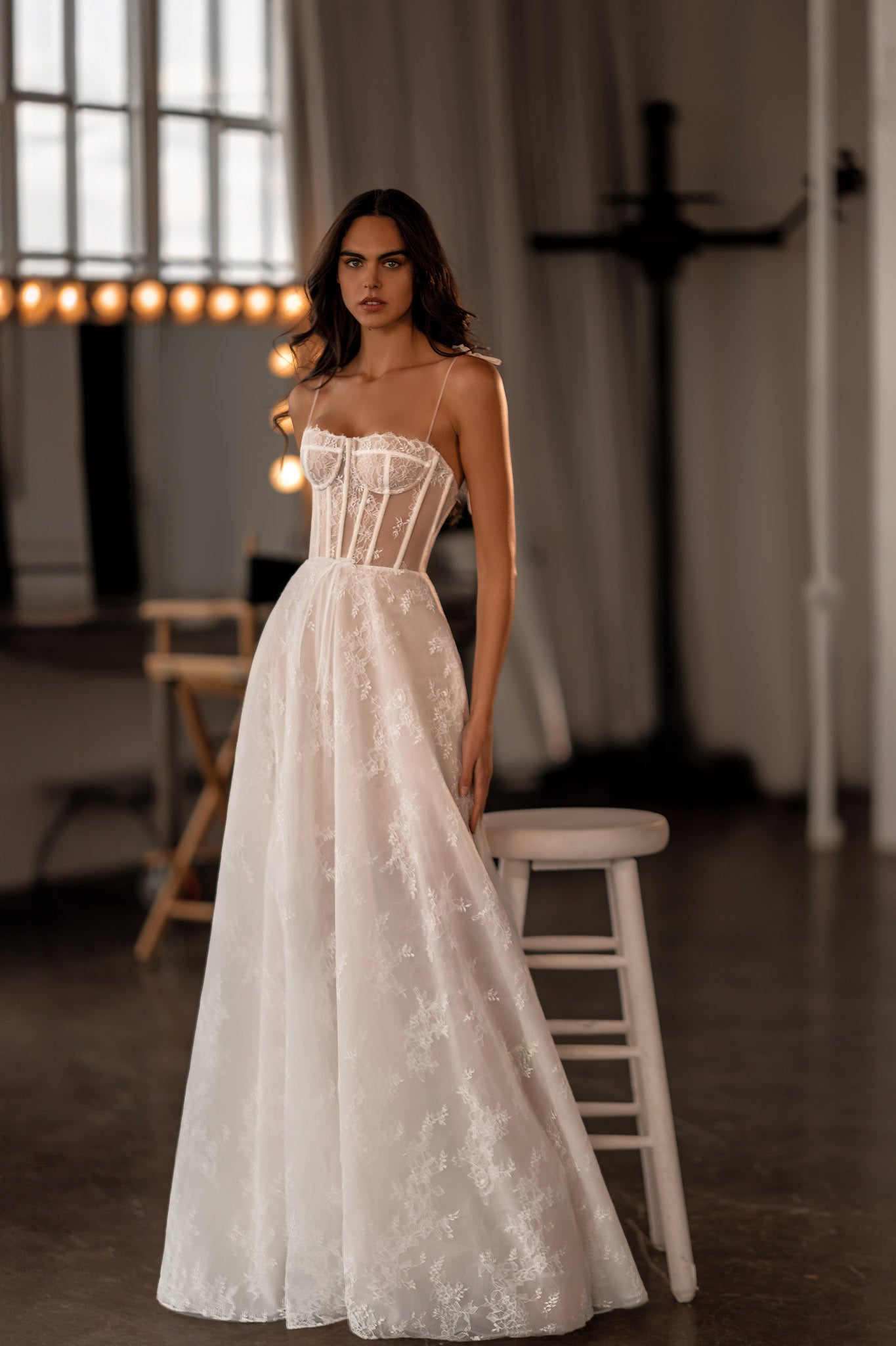 A woman wearing a white, floral lace wedding dress with a sweetheart neckline and sleeveless bodice, standing in a spacious room with high ceilings.